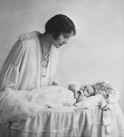 A young woman in a loose pale dress and long strings of pearls, dark hair cropped short, leans over a frilled cot and looks down at the baby lying in it, in black and white.
