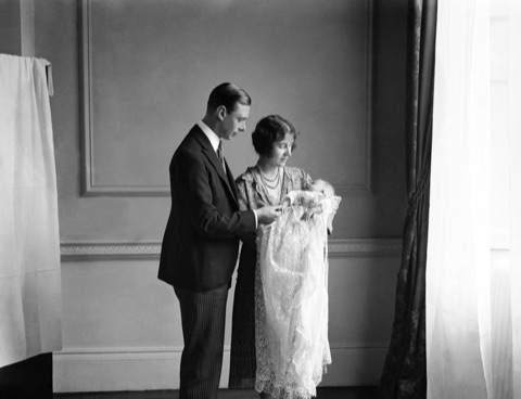 A man in a dark suit and a woman in a patterned dress stand in a panelled room beside a tall curtained window; she holds a baby in a long lace christening robe while he looks down at the child, in black and white.