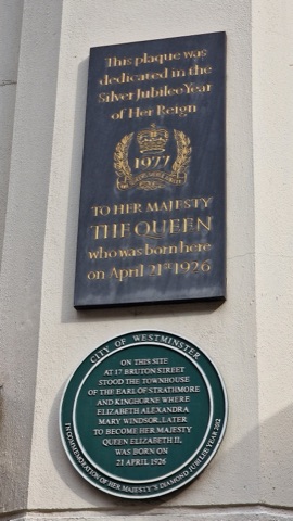 Two plaques on a pale stone wall: a black rectangular one with gold lettering, dedicated in 1977 to the Queen who was born here on 21 April 1926, and a round green City of Westminster plaque below it recording the Earl of Strathmore's townhouse on this site.