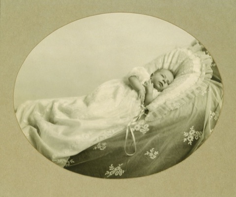 A sepia photograph in an oval vignette of a five-week-old baby lying on a frilled pillow in a long white christening gown that trails over an embroidered coverlet, eyes open, looking at the camera.