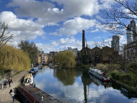 The Regent's Canal on a bright spring day: narrowboats moored along both banks, a willow in new leaf, a lock and a footbridge in the distance, an old brick chimney and warehouse on the right and glass towers beyond, with people walking the towpath.