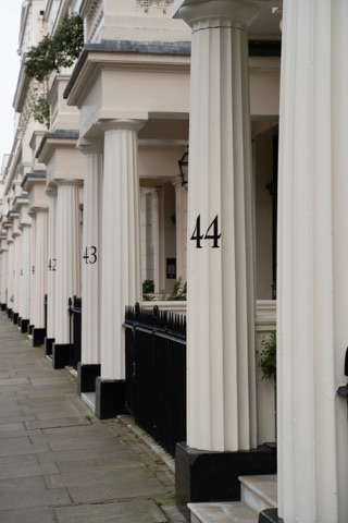 A row of cream stucco porches with fluted Doric columns receding down a Belgravia terrace, the house numbers 42, 43 and 44 painted in black on the columns, black iron railings between them and a paved pavement alongside.