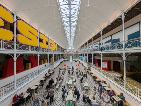 A long museum hall under a glazed barrel roof, white iron columns and galleries down both sides, families at tables across the patterned floor.
