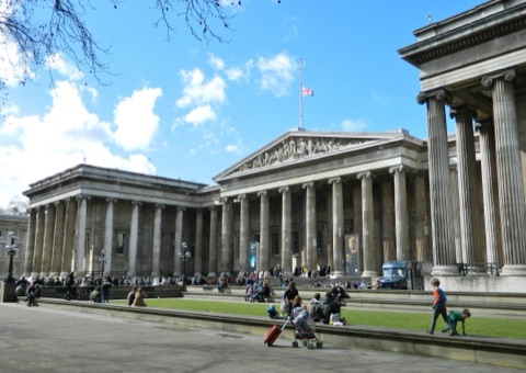 The British Museum's front from its forecourt: a long row of Ionic columns under a sculpted pediment, a flag at half mast, and people sitting on the steps and along the lawn under a blue sky.