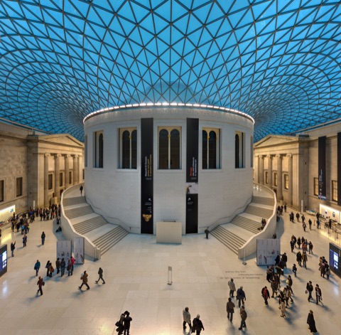 The Great Court of the British Museum from above: the round Reading Room in the middle under a glass roof of triangular panes, curved staircases either side and people crossing the white floor.