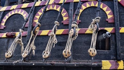The black hull of a replica galleon with red and yellow painted arches, its rigging tied off through wooden blocks, and a cannon's muzzle at a gunport.