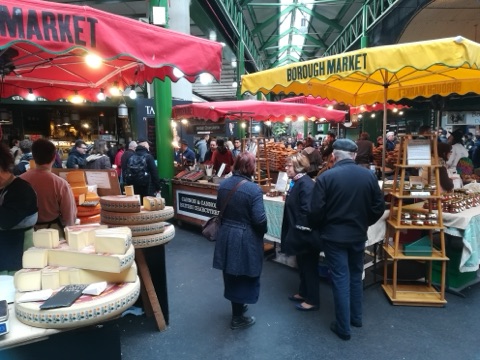 Borough Market under its green iron roof: wheels of cheese and cut wedges on a stall in front, red and yellow awnings lettered with the market's name, a charcuterie counter and shoppers in winter coats between the stalls.
