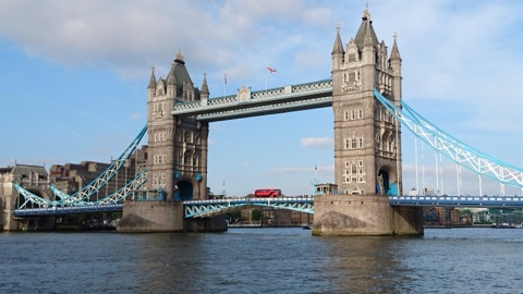 Tower Bridge from the south bank: the two stone towers, the high walkways and the pale blue chains, a red bus crossing, under a blue sky with light cloud.