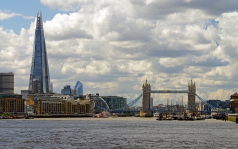 The south bank from the river: the brick warehouses of Butler's Wharf on the left, the Shard rising behind, City Hall's glass, and Tower Bridge on the right with sailing barges moored in front.