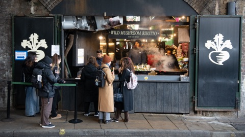 A stall set into a brick railway arch, its green shutters painted with a turnip, a sign reading Wild Mushroom Risotto, steam rising from a wide pan, and a queue of people in coats at the counter.