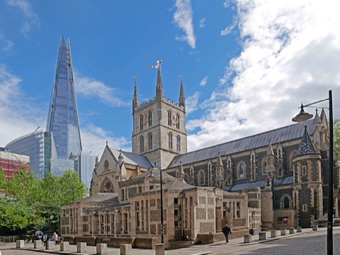 Southwark Cathedral from the north-west: its chequered stone tower and long slate roof, the flint chapter house in front, and the Shard rising behind on a bright day.
