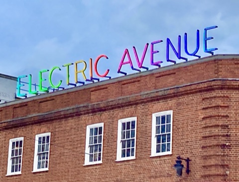 A neon sign reading ELECTRIC AVENUE along the parapet of a red brick corner building, each letter a different colour against a pale sky.