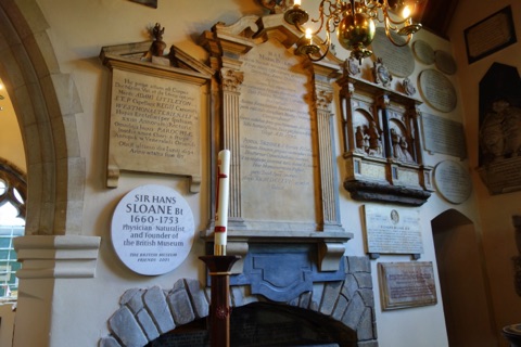 A wall of memorials inside Chelsea Old Church: a round white tablet to Sir Hans Sloane, physician, naturalist and founder of the British Museum, among carved stone monuments, a tall candle in front and a brass chandelier above.