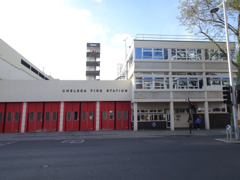 Chelsea Fire Station on the King's Road: a low white 1960s block with a row of red appliance doors, a drill tower behind and a pedestrian crossing in front.