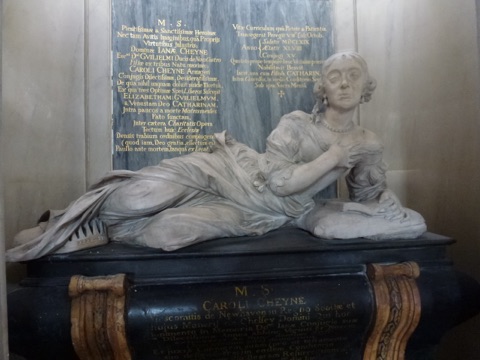 A marble monument of a woman reclining on a cushion in loose robes, one hand on her breast, against a black tablet of gilt Latin inscription, on a dark marble tomb chest.
