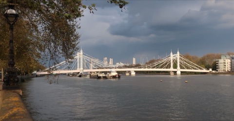 Albert Bridge's white cables and towers across the Thames under a dark storm sky, houseboats moored in front and the towers of the City far behind, seen from under a plane tree on the embankment.