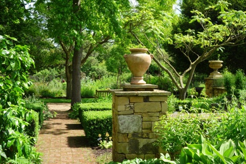 A terracotta urn on a stone pier at the end of a brick path between box hedges, under a spreading tree in the Physic Garden, with a bench beyond.
