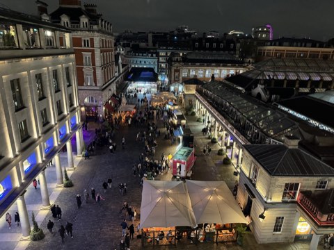 Covent Garden's piazza at night from above, the market building lit and crowds crossing the cobbles