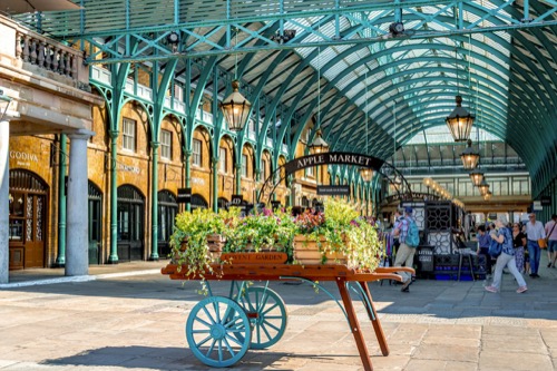 A flower-filled wooden barrow lettered Covent Garden standing on the paving under the market's green iron and glass roof, an Apple Market sign hanging above it and shoppers along the stone arcade beyond.