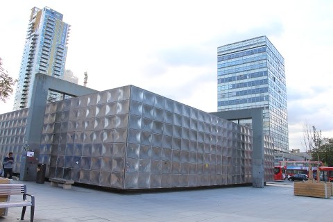 The Michael Faraday Memorial at Elephant and Castle: a low windowless box clad in dulled stainless steel panels, standing on a paved square between concrete portal frames, with a balconied residential tower and a glass office slab rising behind it and a red bus passing on the right.