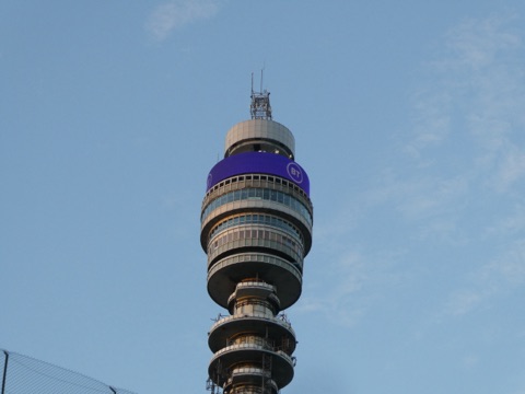 The top of the BT Tower against a pale evening sky: stacked round galleries of glass and the aerials above them, a purple band with the BT logo below the top, seen from street level.