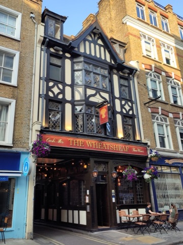 The Wheatsheaf's black-and-white half-timbered front with leaded windows, a hanging sign and baskets of flowers, and two people at a table outside on the pavement.