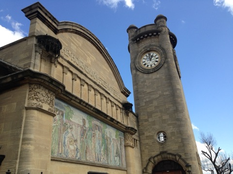 The Horniman Museum from its gardens: a long building in pale stone with a square clock tower at one end, standing above mown grass and planting.
