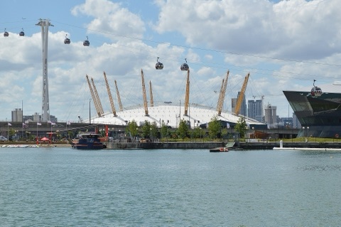 The white dome of The O2 under twelve yellow masts, seen across a dock, with cable car cabins strung on wires in the sky above it.