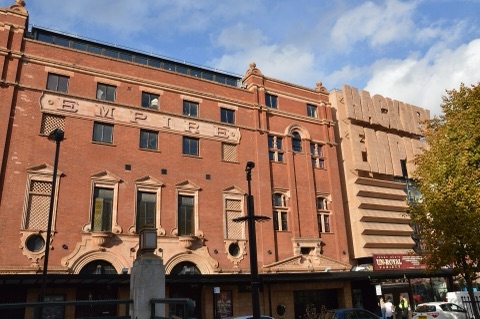 The red brick and terracotta front of a large theatre with EMPIRE carved across it in stone letters, and a modern block-lettered sign standing out beside it.