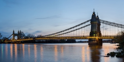 A lit suspension bridge at dusk seen from the bank, two ornate towers rising from the river, the lamps along its deck reflected in the water below.