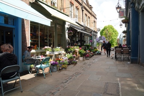 A narrow paved lane between shopfronts, buckets of cut flowers and potted plants down one side, cafe tables out, and people walking away up the hill.