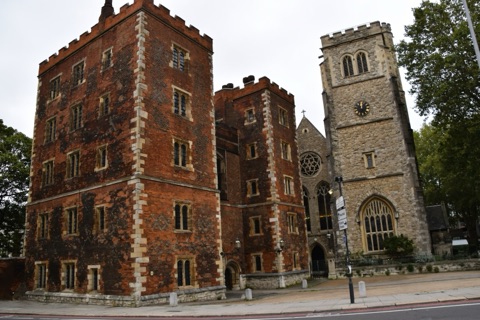 A red brick Tudor gatehouse with two battlemented towers and stone-edged windows, standing at the pavement edge; beside it the pale stone tower of a church, with a clock and a tall traceried window below it
