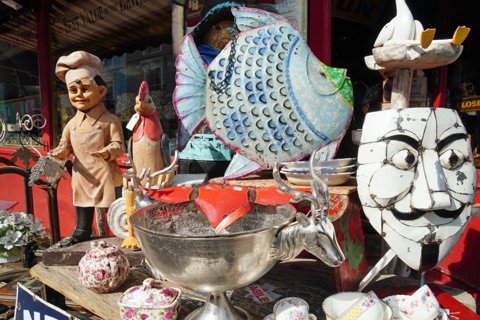 A crowded shopfront on Portobello Road: a painted chef figure, a big tin fish, a white metal mask, a silver bowl with stag's-head handles and flowered china, stacked in front of a red-painted antiques shop.