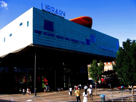 A blue-green box of a building held up on thin steel legs over an open square, LIBRARY in cut letters on its roof and an orange pod projecting from it, under a deep blue sky.