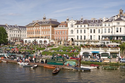 Richmond Riverside on a sunny afternoon: brick and white stucco buildings above terraced lawns crowded with people sitting out, rowing boats and a moored barge along the bank, the Thames in the foreground