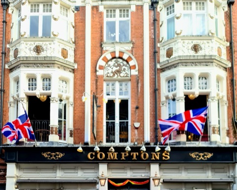 The upper floors of Comptons on Old Compton Street: red brick and cream stone with carved swags and a relief over the middle window, two Union flags flying from the bay windows, gilt lettering on a black fascia and a rainbow flag over the door.