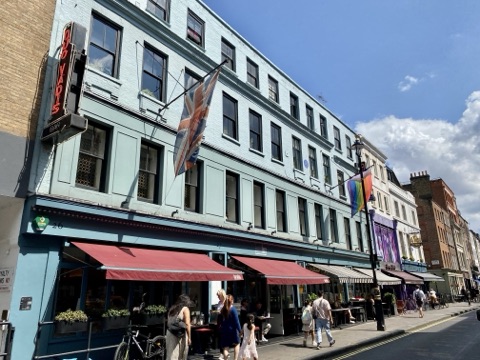 A pale blue Georgian terrace with red awnings over its ground-floor restaurant, a neon 'Quo Vadis' sign on the corner, a Union flag and a rainbow flag, and people at pavement tables in summer sun.