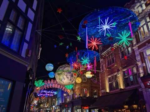 Carnaby Street at night under its Christmas lights: glowing planets, neon stars and blue foil discs strung across the street, with 'Carnaby celebrates' in lit letters over the shops.