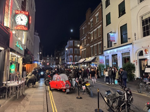 Frith Street at night: motorbikes and a red bubble car parked down the middle of the road, a crowd of riders and drinkers on both pavements, the neon clock of Bar Italia on the left and the Delhi Brasserie's lit front on the right.