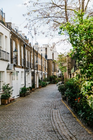 A cobbled mews curving away between low houses: white-painted garages and brick fronts with iron balconies on the left, climbing greenery and pots of shrubs along both sides, and a bare tree overhead.
