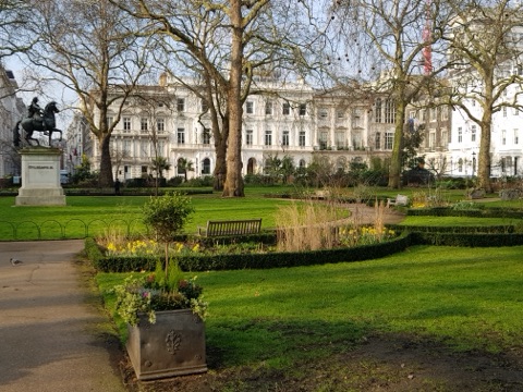 A garden square in early spring: lawn, clipped box hedges and a bench under bare plane trees, an equestrian statue on its plinth at the left, and white stucco terraces behind.