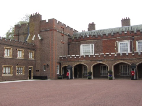 A courtyard of St James's Palace: dark red Tudor brick with battlements and tall chimneys, an arcade of pointed arches along the ground floor, and two guardsmen in red tunics and bearskins at their sentry boxes.