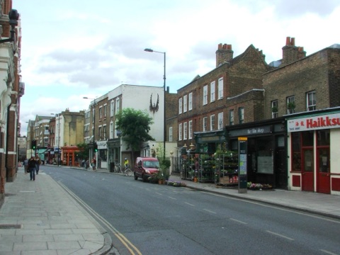 Stoke Newington Church Street: low brick terraces either side of a narrow road, small shopfronts at street level with a florist's racks out on the pavement, a plane tree, and a large phoenix painted on a white gable end.