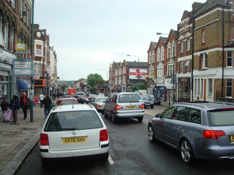 Sydenham Road looking east: a parade of red brick Victorian shops with gabled upper floors along the right-hand side, traffic queueing in both lanes, and a fingerpost on the near pavement pointing to Downham, Catford and Penge.