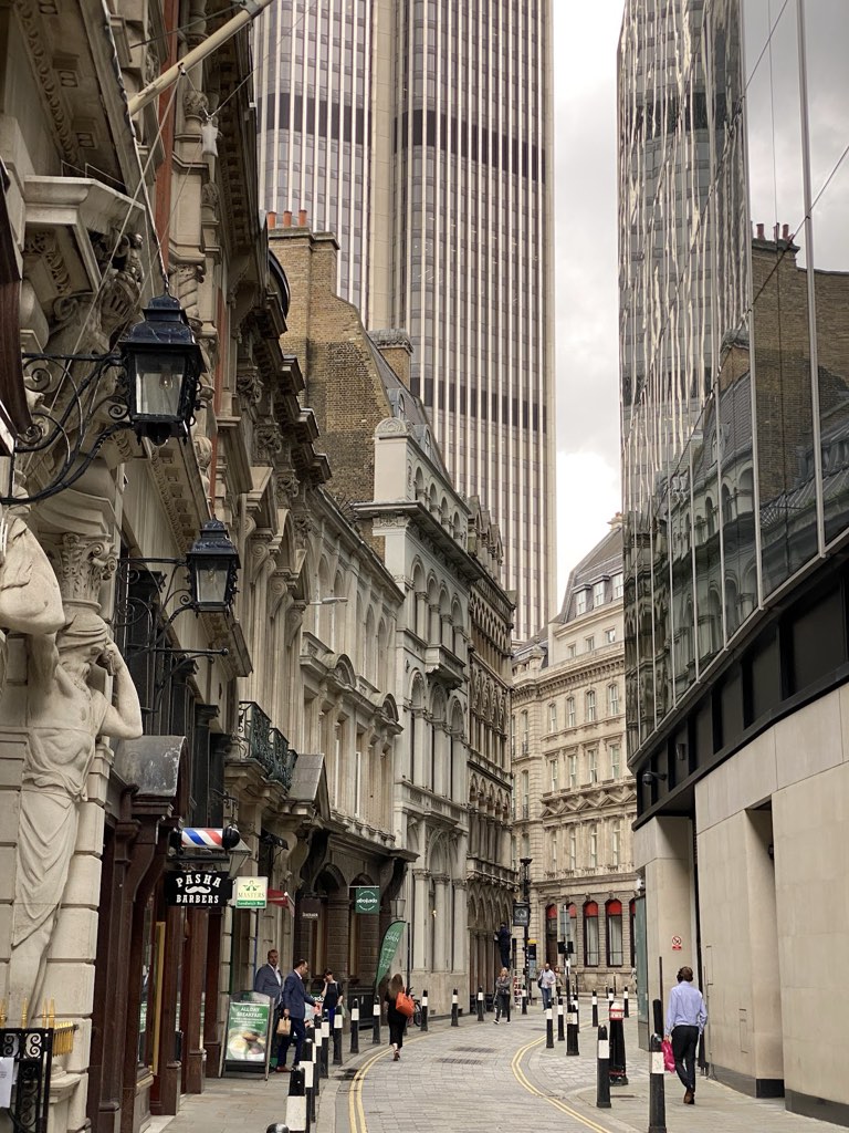 A narrow City lane between stone facades, with glass towers rising directly behind them