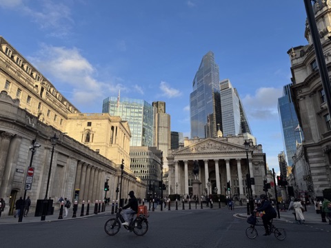 Bank junction: the Royal Exchange's tall columned portico and carved pediment, with the equestrian statue of Wellington in front, the Bank of England's stone wall on the left and the glass towers of 22 Bishopsgate rising behind, on a bright afternoon with cyclists crossing.