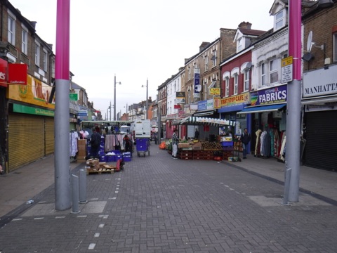 Walthamstow High Street market: a pedestrianised street of stalls under awnings, fruit and vegetables and clothing racks in front of shopfronts, running away into the distance.
