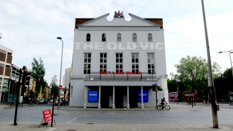 The Old Vic's pale stucco front on a street corner, its name in raised letters across the upper storey and 'We'll be back' in red on the balcony, a cyclist passing on an empty road and red buses beyond.