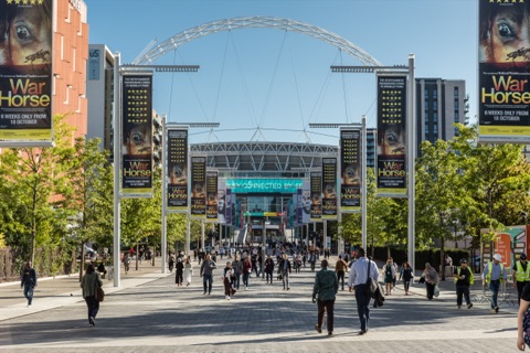 Crowds walking up Olympic Way towards Wembley Stadium on a bright afternoon, the arch above the roof and banners hanging from the lamp posts