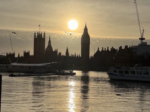 The Palace of Westminster and the clock tower in silhouette against a low sun, seen across the Thames from the South Bank, with a pier and a moored boat in front, gulls in the air and light on the water.